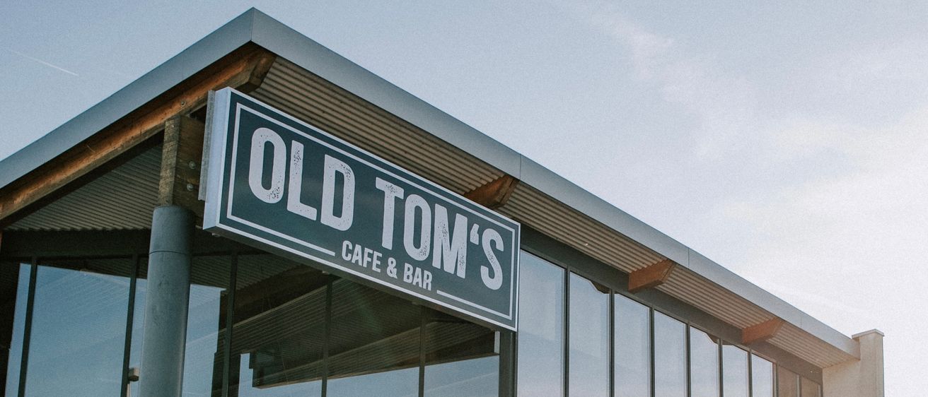 Eine Gruppe von Menschen sitzt an Tischen vor dem Old Tom's Café und Bar, das sich in einem modernen Gebäude mit Glaswänden und einem großen Schild über dem Eingang befindet.