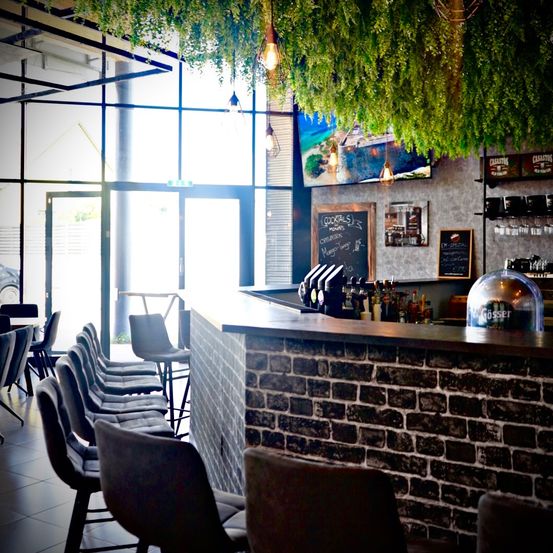 Innenansicht eines Cafés mit einer großen Tafel, auf der Cocktails und Getränke stehen. Das Café hat eine Ziegelmauer und eine Theke mit Flaschen und Gläsern.