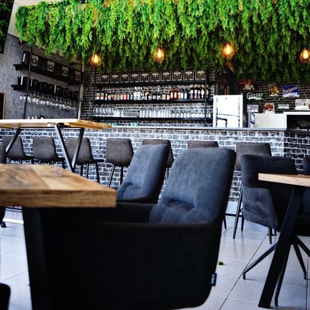 Eine Bartheke mit Regalen, die Flaschen und Weingläser halten, beleuchtet von hängenden Lampen. Grüne hängende Pflanzen schmücken die Decke. Leere Tische und Stühle umgeben die Theke.