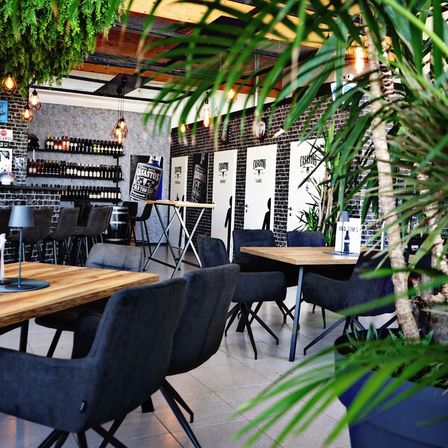 Ein Innenrestaurant mit Tischen, Stühlen und Topfpflanzen. Es gibt hängende Lampen und ein Regal mit Flaschen.