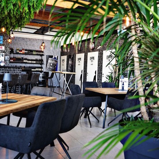 Ein Innenrestaurant mit Tischen, Stühlen und Topfpflanzen. Es gibt hängende Lampen und ein Regal mit Flaschen.