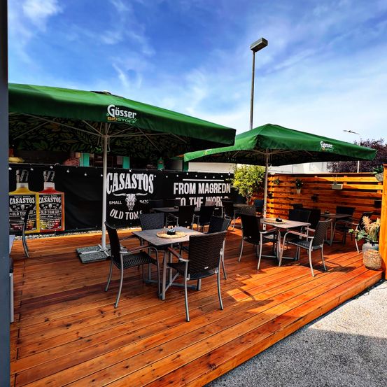 Terrassenbereich mit Holzboden, Tischen und Stühlern sowie grünen Sonnenschirmen. Dahinter befindet sich eine Holzwand mit Pflanzen und einem Schild.