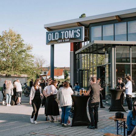 Eine Gruppe von Menschen steht vor dem Old Tom's Cafe & Bar und unterhält sich. Die Einrichtung hat Glaswände und ein Schild über dem Eingang.