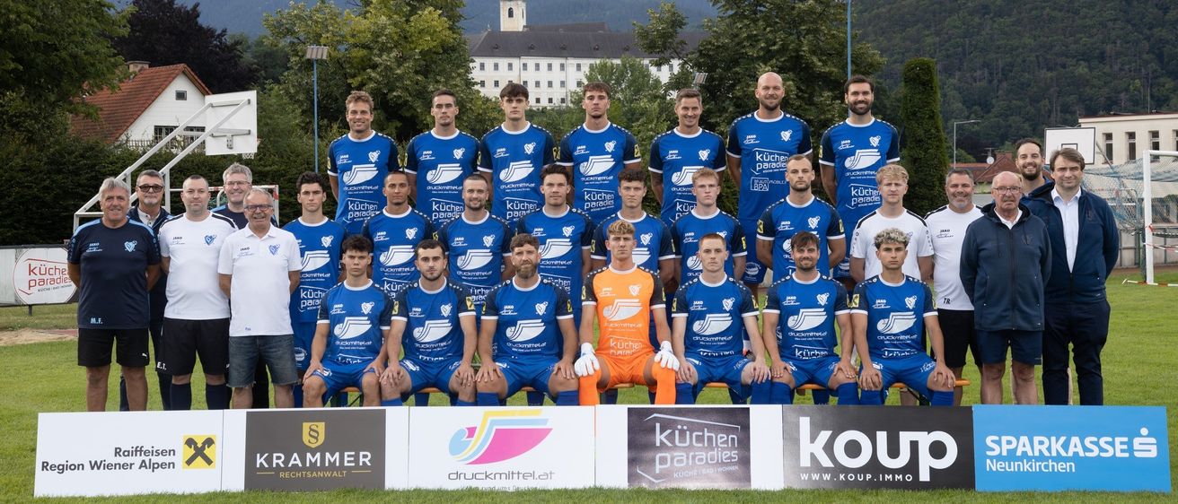 Ein Fußballteam posiert für ein Foto auf einem Feld, mit einem Banner, das 'KRAMMER' im Hintergrund bewirbt. Sie tragen blaue Uniformen mit weißen Akzenten. Ein Torwart in Orange ist in der vorderen Reihe.