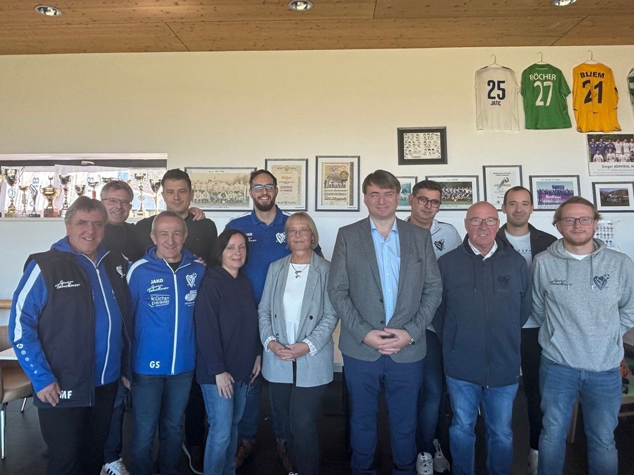 Eine Gruppe von Menschen in Jacken, darunter zwei Frauen, steht vor einer Wand mit gerahmten Bildern und Trikots. Der Mann in der Mitte trägt einen Anzug und alle schauen zur Kamera.