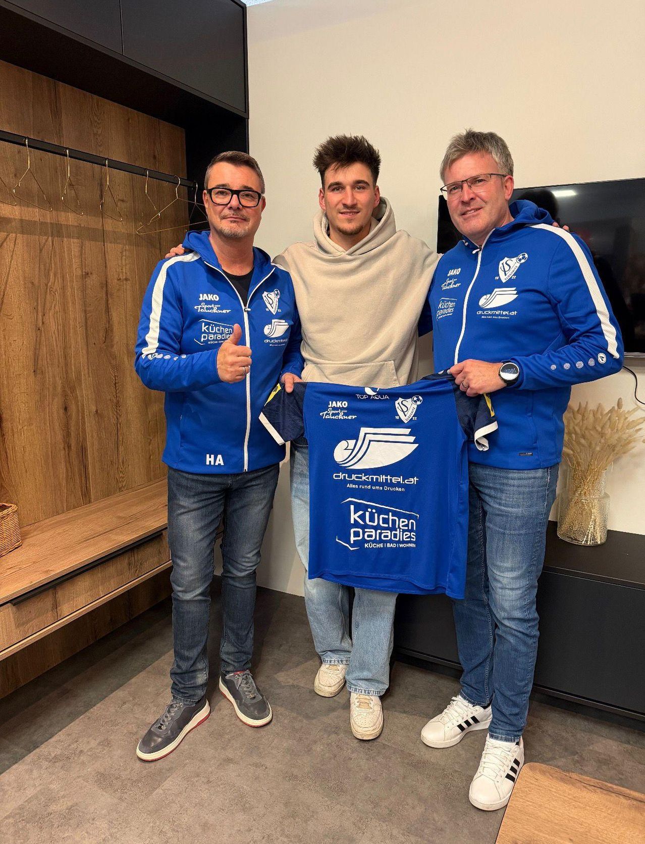 Drei Männer in blauen Jacken stehen zusammen, einer gibt einen Daumen hoch und hält ein blaues Shirt mit dem Text 'Kuchen Paradies'. Sie befinden sich in einem Raum mit einer Holzwand und einem Fernseher.