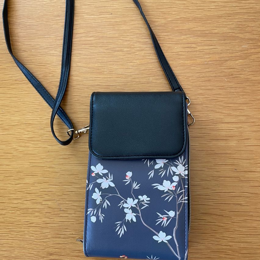 Eine schwarze Umhängetasche mit Blumenmuster auf einer Holzoberfläche. Die Tasche hat einen Reißverschluss und einen abnehmbaren Riemen.
