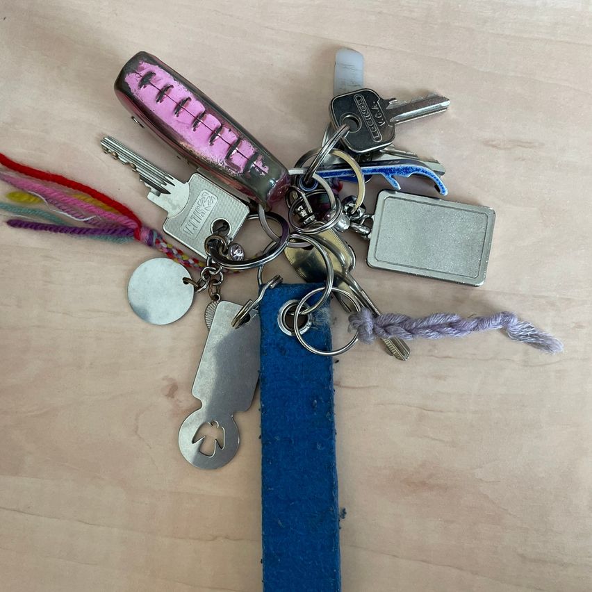 Eine Sammlung von Schlüsseln an einem blauen Band, einschließlich eines rosa Schlüssels mit Kratzern, eines silbernen Schlüssels und eines silbernen Anhängers. Der Hintergrund ist hölzern.