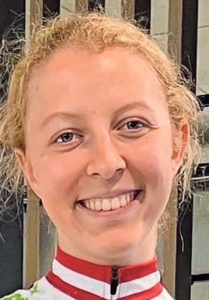 Eine Frau mit kurzen blonden Haaren lächelt breit und zeigt ihre Zähne. Sie trägt einen roten Kragen und scheint sich drinnen zu befinden.