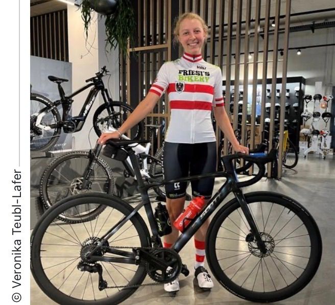 Eine Frau in einem Radtrikot steht neben einem Fahrrad in einer Ausstellung. Das Fahrrad hat eine Wasserflasche daran befestigt. Hinter ihr sind verschiedene Fahrräder ausgestellt.