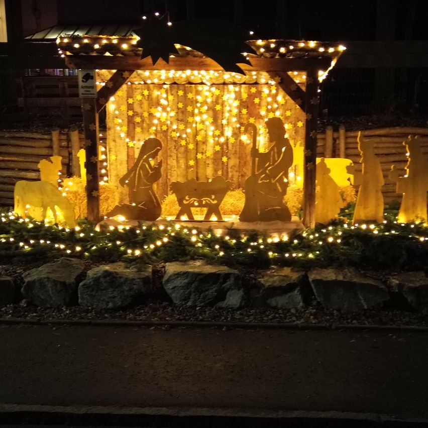 Eine Krippe mit Maria und Josef, einem Baby und Tieren, umgeben von leuchtenden Lichtern und Holzzäunen in der Nacht.