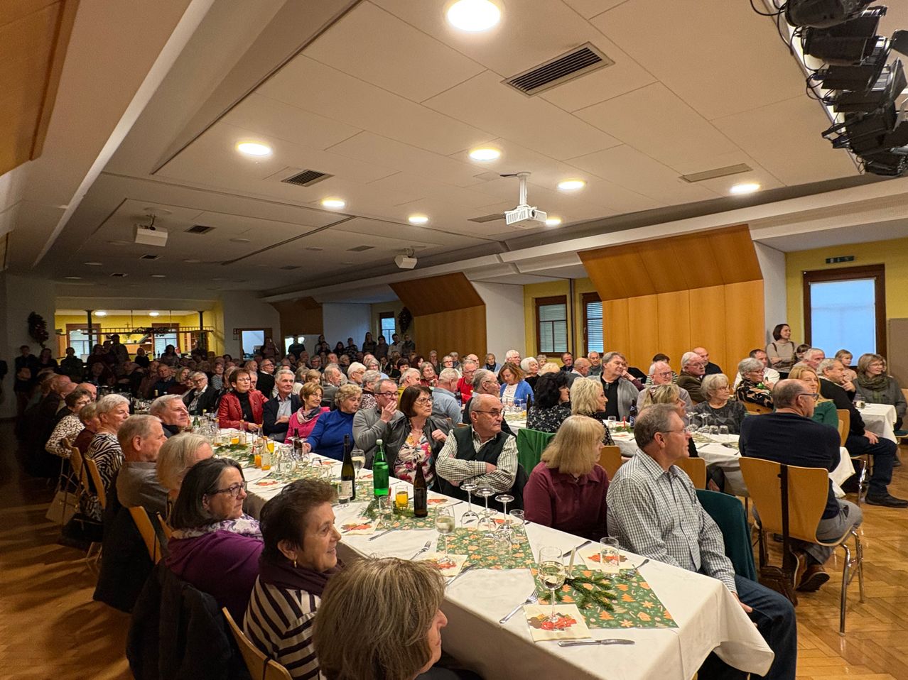 Ein Saal mit vielen Menschen, die an langen Tischen sitzen, einige klatschen. Auf den Tischen stehen Gläser, Flaschen und Blumen. Der Saal hat Holzwände und weiße Decken mit Lampen.