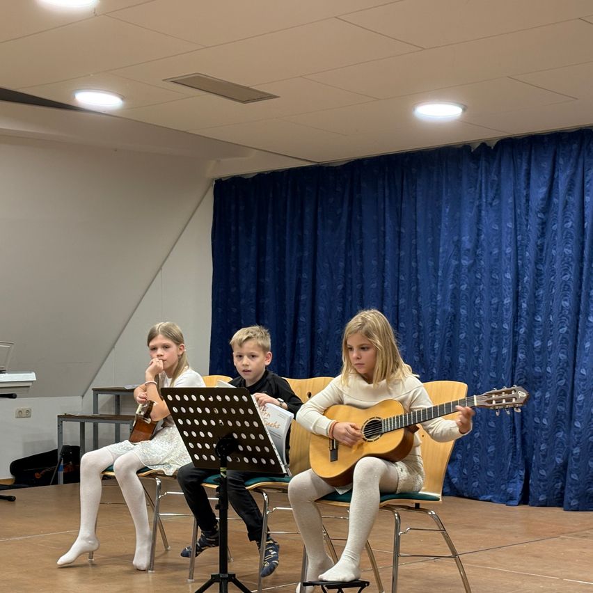 Drei Kinder sitzen auf Stühlen und spielen Gitarre. Ein Mädchen auf der rechten Seite hält eine Gitarre und scheint zu spielen. Das Mädchen auf der linken Seite hält eine Gitarre und schaut auf das Notenblatt. Der Junge in der Mitte schaut auf das Mädchen auf der linken Seite. Hinter ihnen ist ein blauer Vorhang.