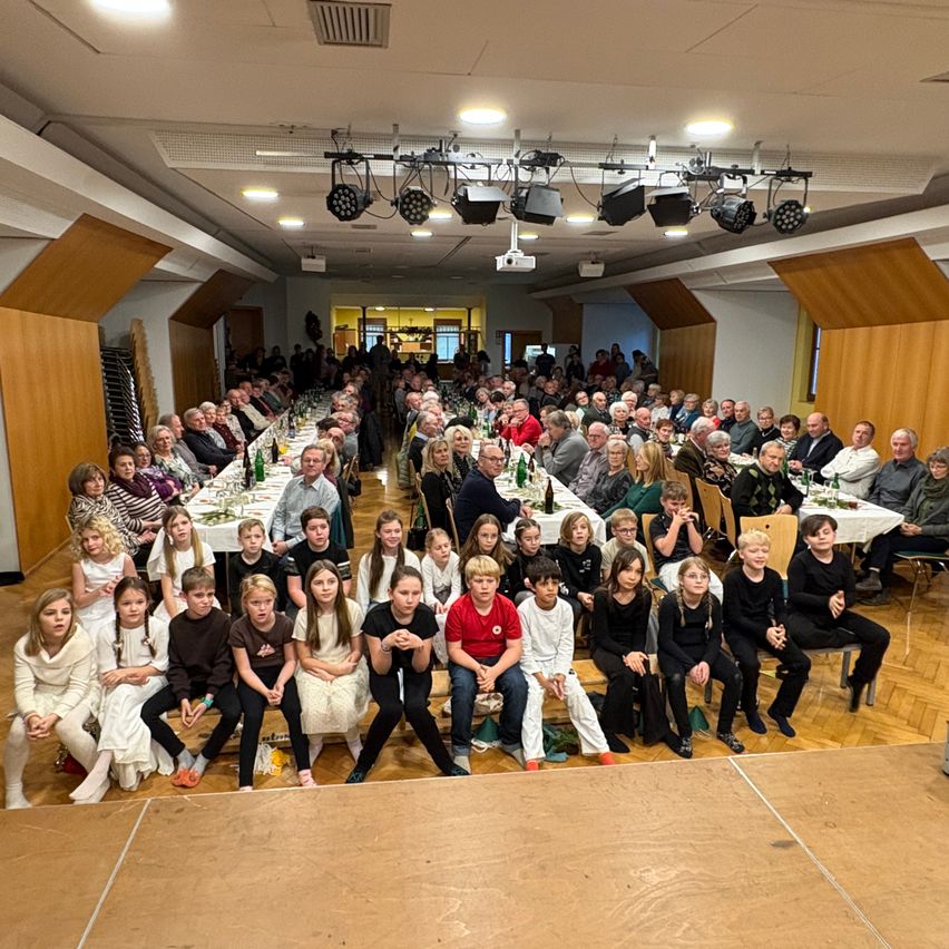 Eine große Gruppe von Menschen, darunter Kinder und Erwachsene, sitzen an langen Tischen in einem Bankettsaal. Der Raum ist gut beleuchtet, mit Scheinwerfern und Lampen, die von der Decke hängen. Einige Personen stehen hinter den Tischen, möglicherweise beim Servieren von Speisen.