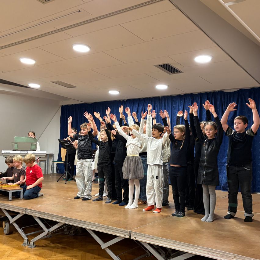 Kinder auf einer Bühne in einer Schule, einige mit erhobenen Händen, während andere auf dem Boden sitzen und Instrumente spielen. Dahinter ein blauer Vorhang und ein Notenständer.