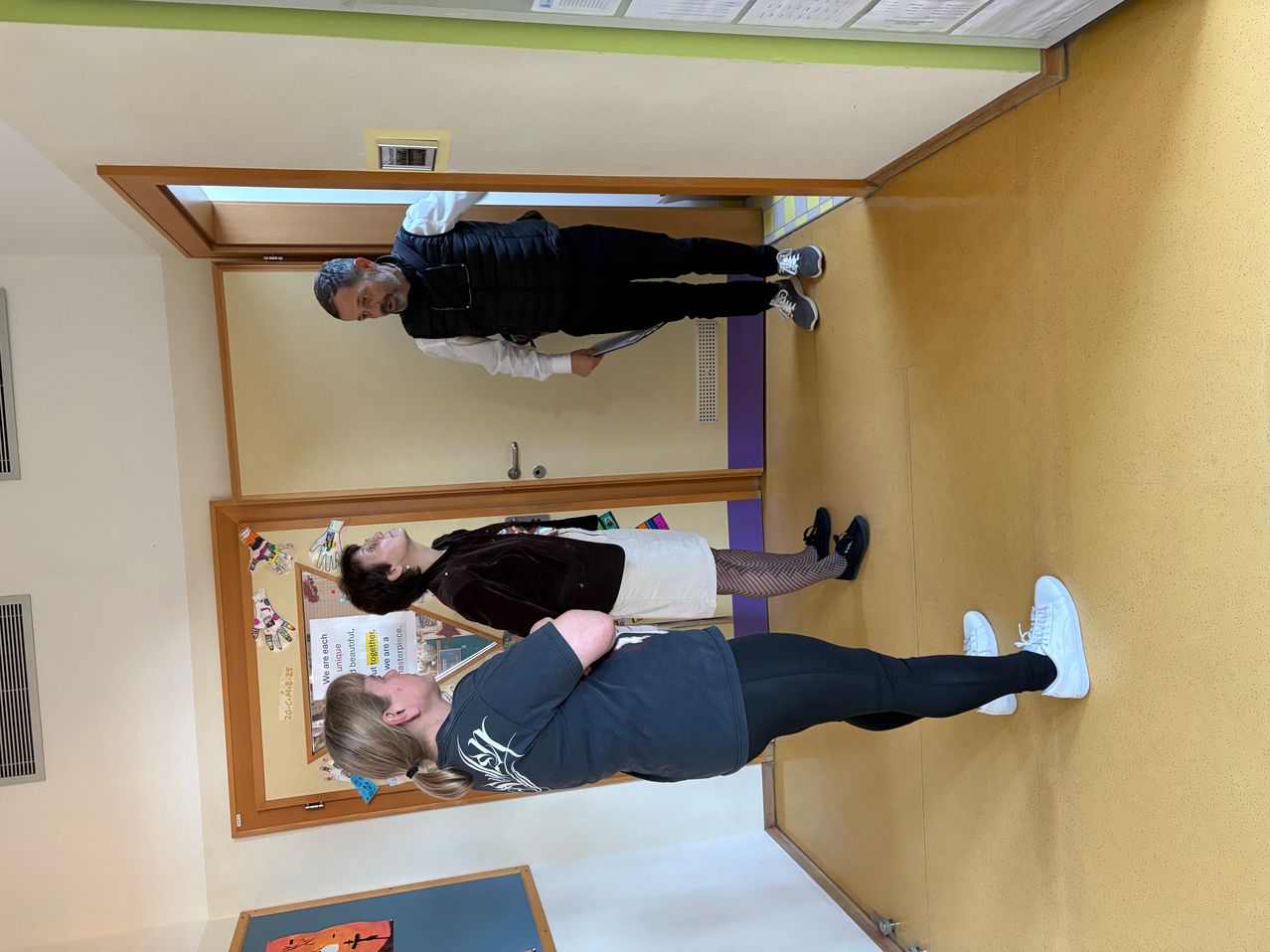 Drei Personen befinden sich in einem Flur, zwei lehnen an der Wand und eine steht. Die Person in der Mitte scheint sich zu strecken oder eine körperliche Aktivität durchzuführen.