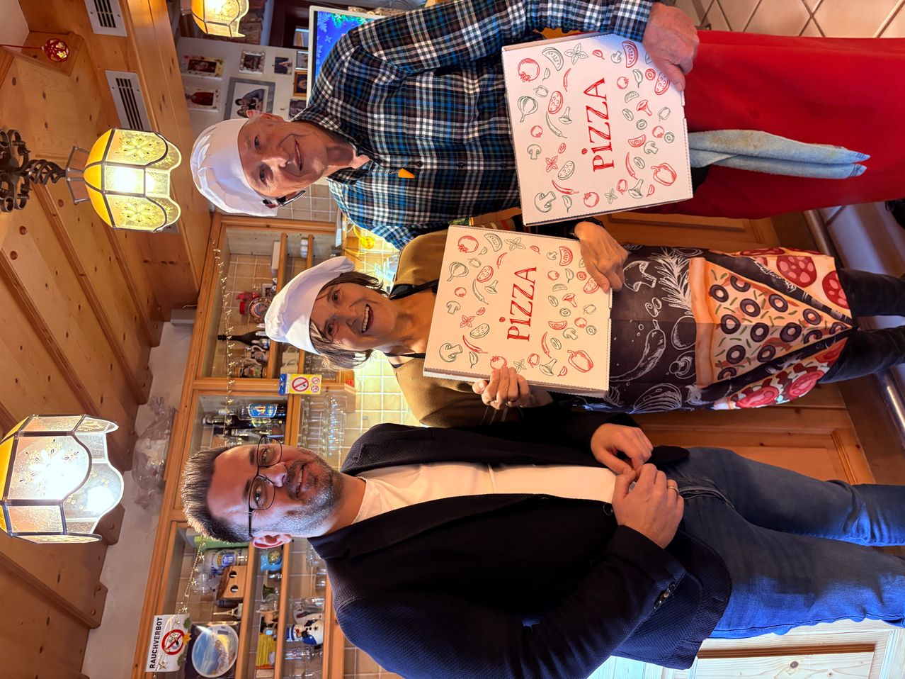 Drei Personen lächeln und posieren für ein Foto in einem Raum. Zwei von ihnen halten Boxen mit dem Wort Pizza. Der Mann auf der rechten Seite trägt eine Kochmütze und ein kariertes Hemd.
