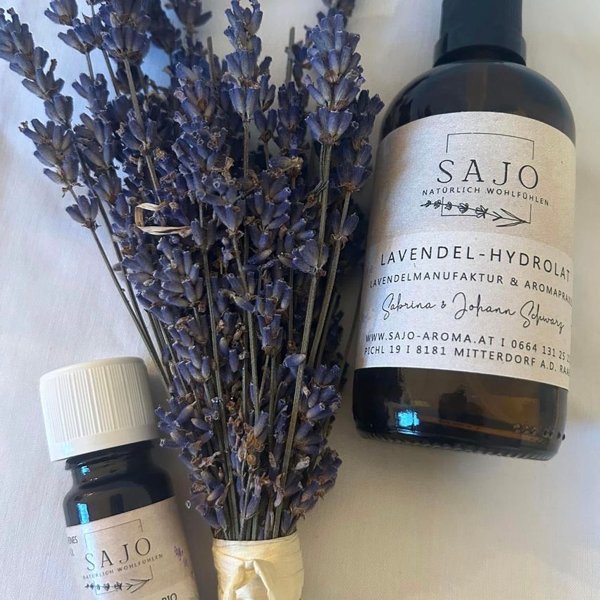 Ein Strauß Lavendelblüten und zwei Flaschen Lavendelhydrolat und ätherisches Öl von SAJO sind auf einem weißen Tuch zu sehen.