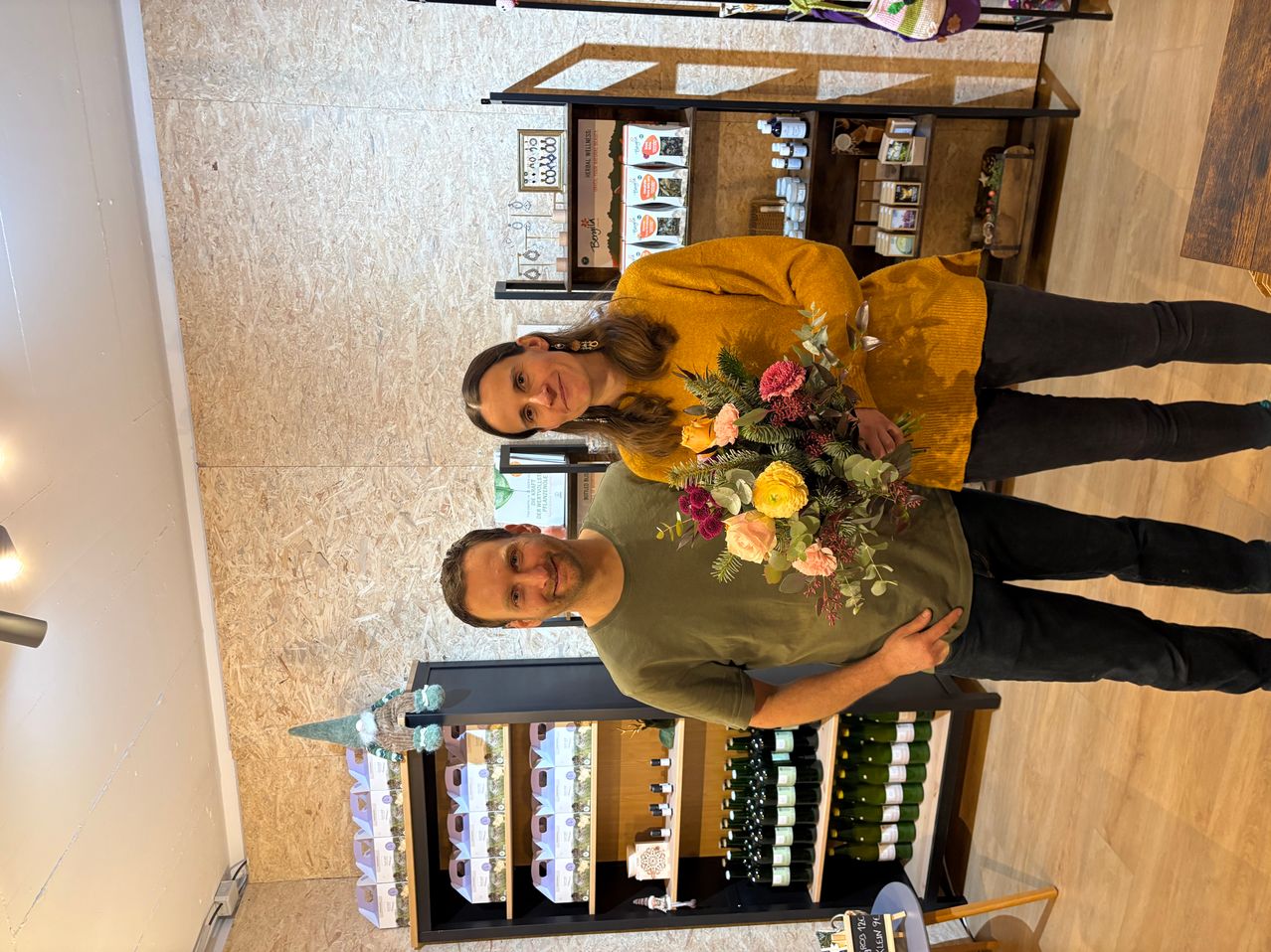 Ein Paar steht in einem Geschäft und hält einen Blumenstrauß. Die Frau trägt einen gelben Pullover und der Mann ein grünes Hemd. Hinter ihnen sind mit Produkten gefüllte Regale zu sehen.