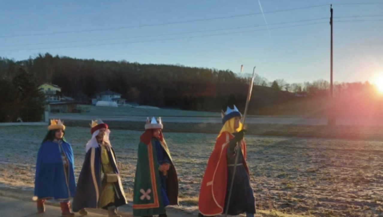 Drei Menschen, gekleidet wie mittelalterliche Könige oder Königinnen, gehen auf einem Feld mit trockenem Gras. Sie tragen Kronen und Umhänge, einer hält einen Stab. Ein Hügel und ein Haus sind im Hintergrund zu sehen.