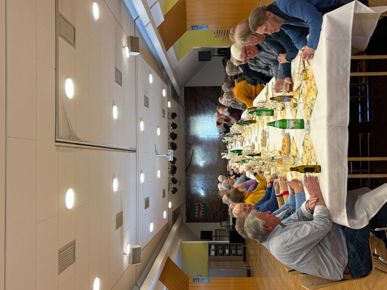 Ein langer Tisch mit vielen sitzenden Erwachsenen, möglicherweise bei einem Bankett, gefüllt mit Weingläsern und Flaschen. Der Raum hat weiße Wände und Deckenleuchten.