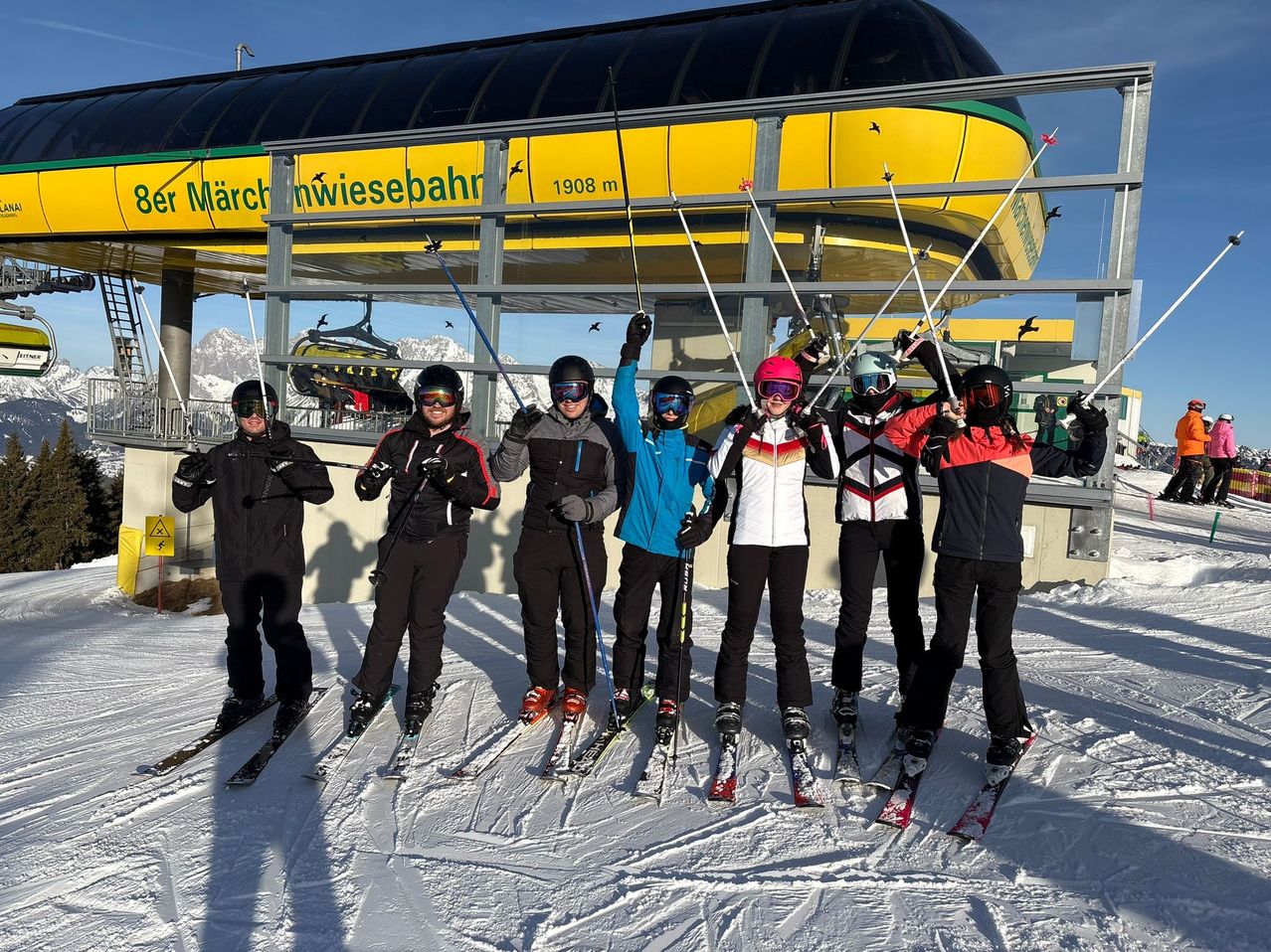 Eine Gruppe von Skifahrern posiert für ein Foto auf einer verschneiten Piste. Sie halten Skistöcke in der Hand und tragen Helme. Hinter ihnen steht eine gelbe Struktur mit den Aufschriften 'Märchenwiesebahn' und '1908 m'. In der Ferne sind Berge zu sehen.