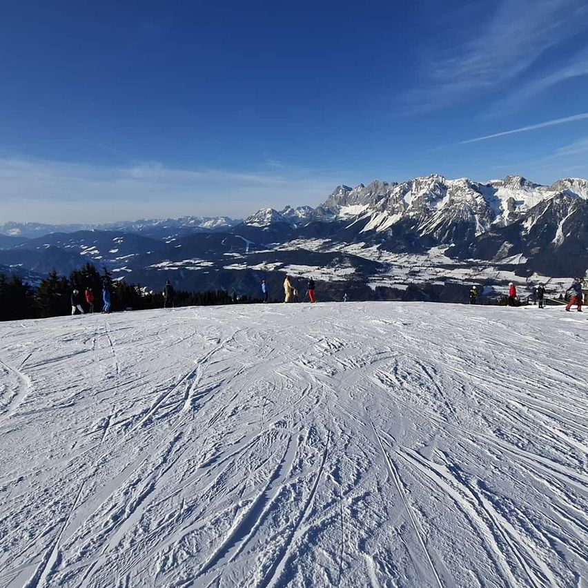 Eine verschneite Berglandschaft mit Menschen, die auf dem schneebedeckten Boden stehen. In der Ferne befinden sich Berggipfel und ein blauer Himmel mit einigen Wolken.