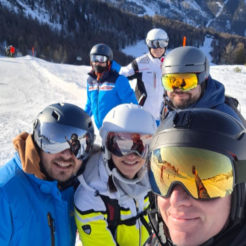 Eine Gruppe von Skifahrern in verschiedenen Farben der Ausrüstung, einschließlich Blau, Gelb und Weiß, stehen auf einem verschneiten Hang. Sie tragen alle Helme und Schutzbrillen. Im Hintergrund befinden sich Bäume und eine Bergkette.