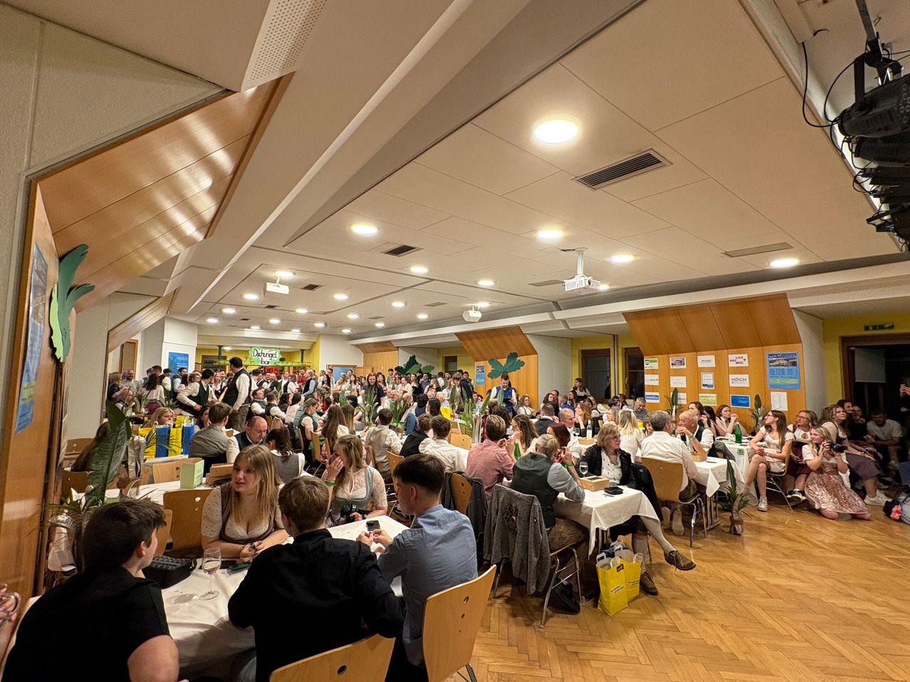 Eine große Menschenmenge, die um Tische in einem Bankettsaal herumsitzt, einige unterhalten sich, andere genießen ihre Mahlzeiten, mit einem Projektor und dekorativen Pflanzen im Hintergrund.