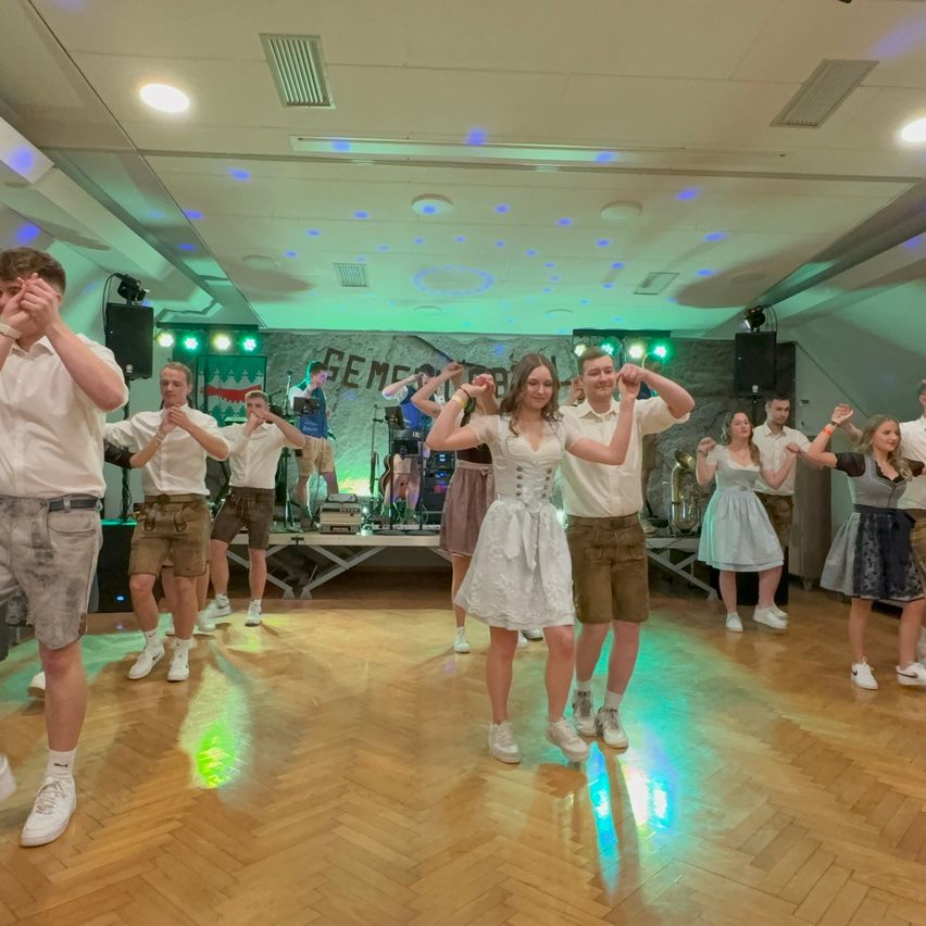 Eine Gruppe von Menschen tanzt auf einem Holzboden in einem Raum mit Bühne und Musikinstrumenten. Einige tragen traditionelle Kleidung.