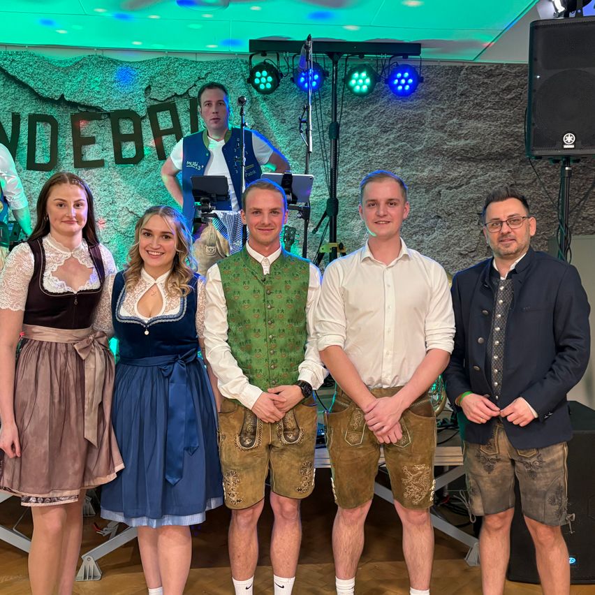 Eine Gruppe junger Menschen in traditioneller deutscher Kleidung posiert für ein Foto auf einer Bühne mit einem Lautsprecher und Musikinstrumenten im Hintergrund.