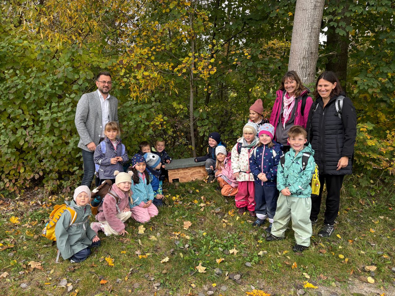 Eine Gruppe von Kindern und Erwachsenen steht und sitzt auf einem Grasfeld. Sie alle lächeln und scheinen für ein Foto zu posieren. Die Kinder tragen Winterkleidung. Vor ihnen steht eine Holzkiste auf dem Boden. Hinter ihnen befinden sich Bäume mit gelben Blättern.