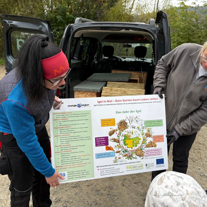 Zwei Frauen halten ein Plakat mit einer Zeichnung eines Regenwurms in der Nähe eines Vans mit offenen Hecktüren, mit Holzkisten im Inneren.