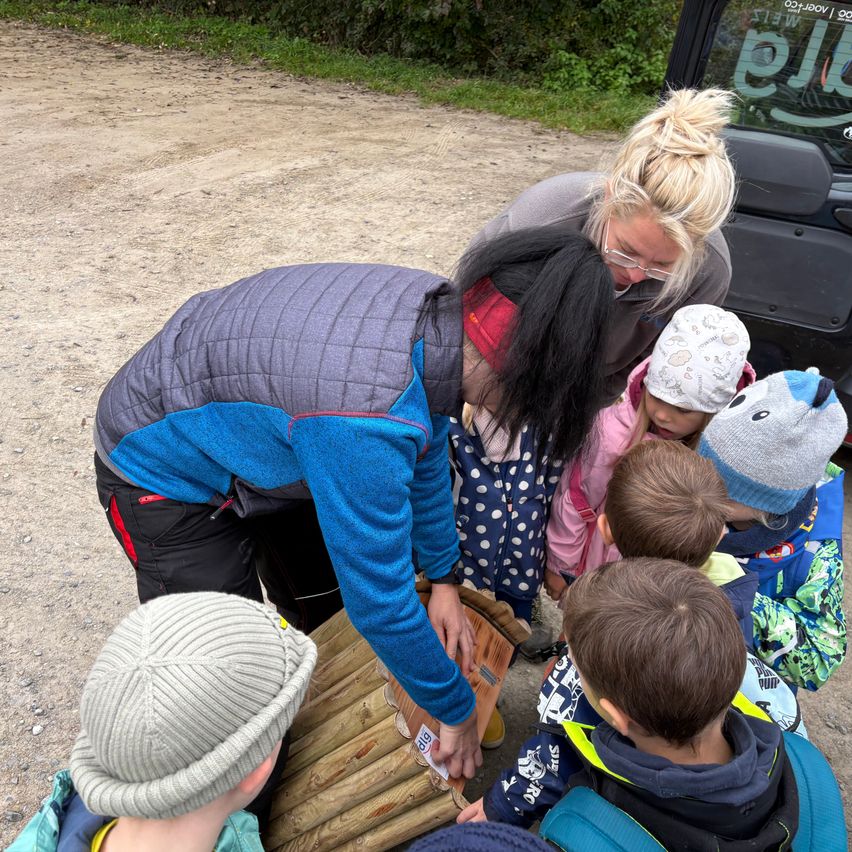 Eine Gruppe von Kindern und einem Erwachsenen sind um ein Holzobjekt versammelt. Der Erwachsene hält das Objekt und erklärt den Kindern etwas. Die Kinder tragen Winterkleidung und Mützen. Im Hintergrund steht ein Fahrzeug mit einem verschwommenen Amazon-Logo.