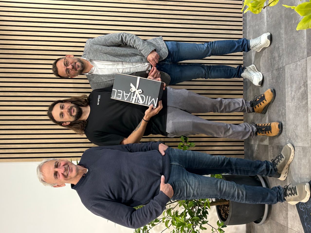 Three men standing together, smiling for a photo. The middle man holds a gift box. They are in front of a wooden wall with a potted plant.