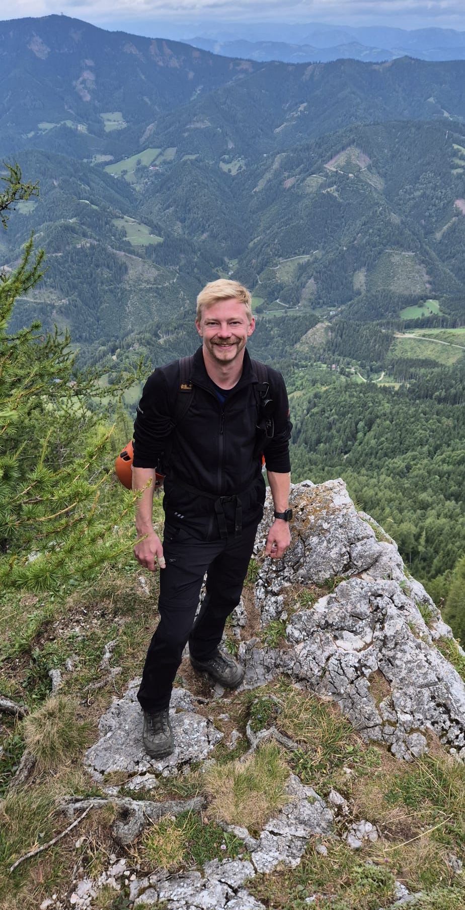 A man with a backpack stands on a mountain peak, smiling and looking forward. Behind him, a forest with green trees and a distant valley.