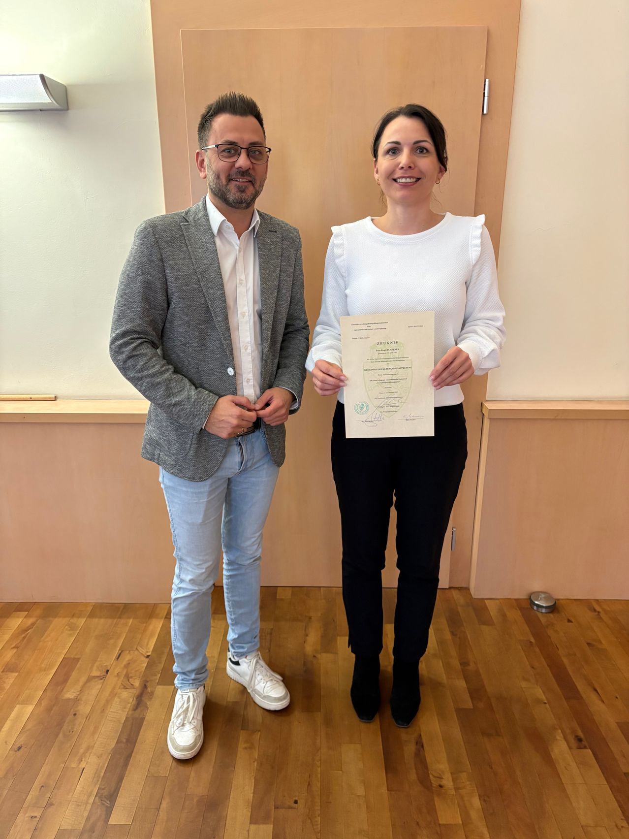 A man and a woman stand next to each other holding a certificate. They are both smiling. The man wears glasses, a gray blazer, and light blue jeans. The woman wears a white blouse, black pants, and black boots. They are in a room with a wooden floor.