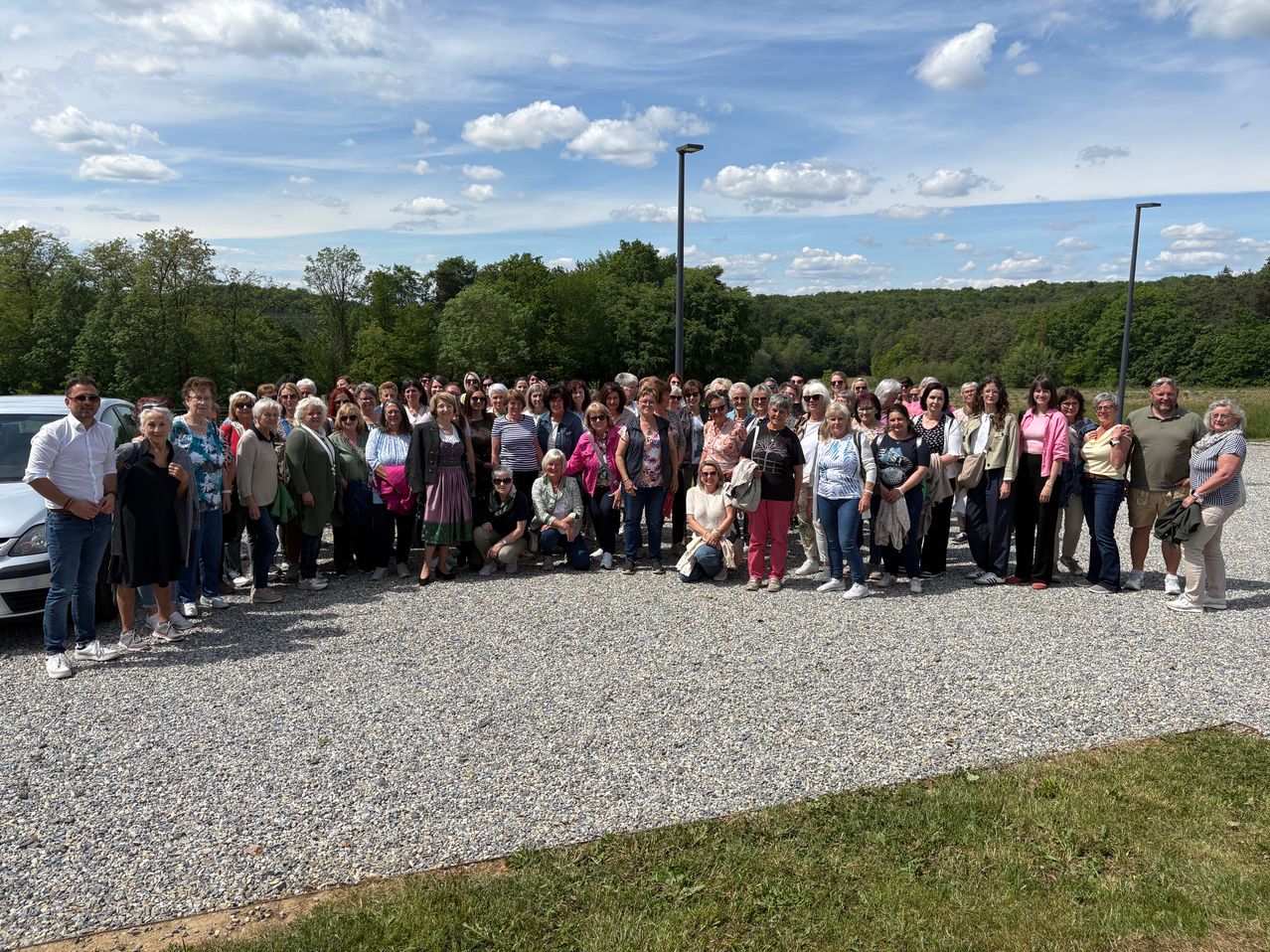A large group of people, mostly women, pose for a photo on a gravel road under a partly cloudy sky with trees in the background.