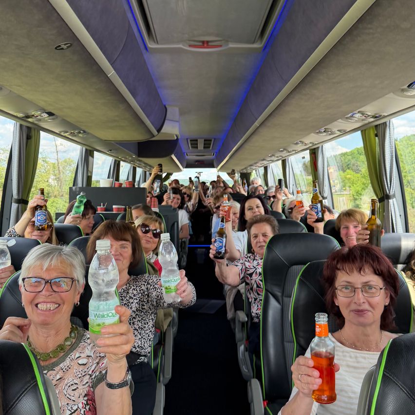 A group of people on a bus, all holding up bottles of beer and water, seem to be on a road trip or tour.