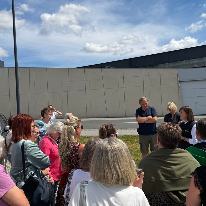 A group of people gathered outdoors, some standing, some sitting, in front of a modern building under a cloudy sky.