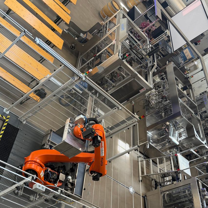 An overhead view of a large, industrial facility with various machines and robots. The robots are connected to a network of cables and have various mechanical arms. The facility has metal structures and wooden benches.