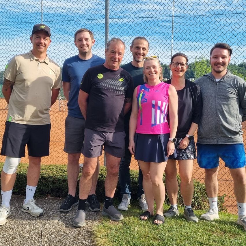 A group of people dressed in casual clothes stand on the grass, likely posing for a photograph on a tennis court.