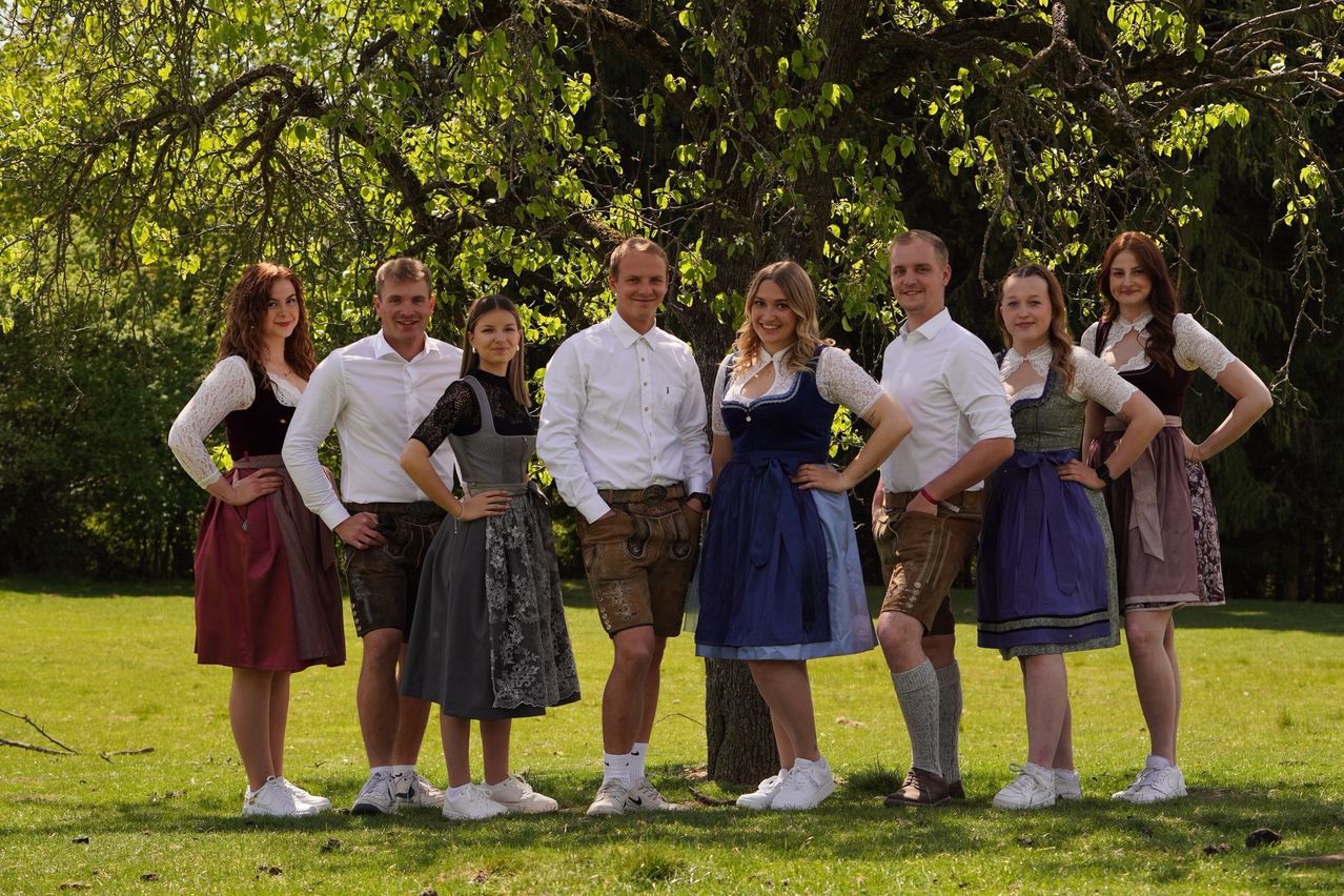 Eine Gruppe von Menschen in traditioneller bayerischer Kleidung posiert für ein Foto in einem Park mit Bäumen im Hintergrund.