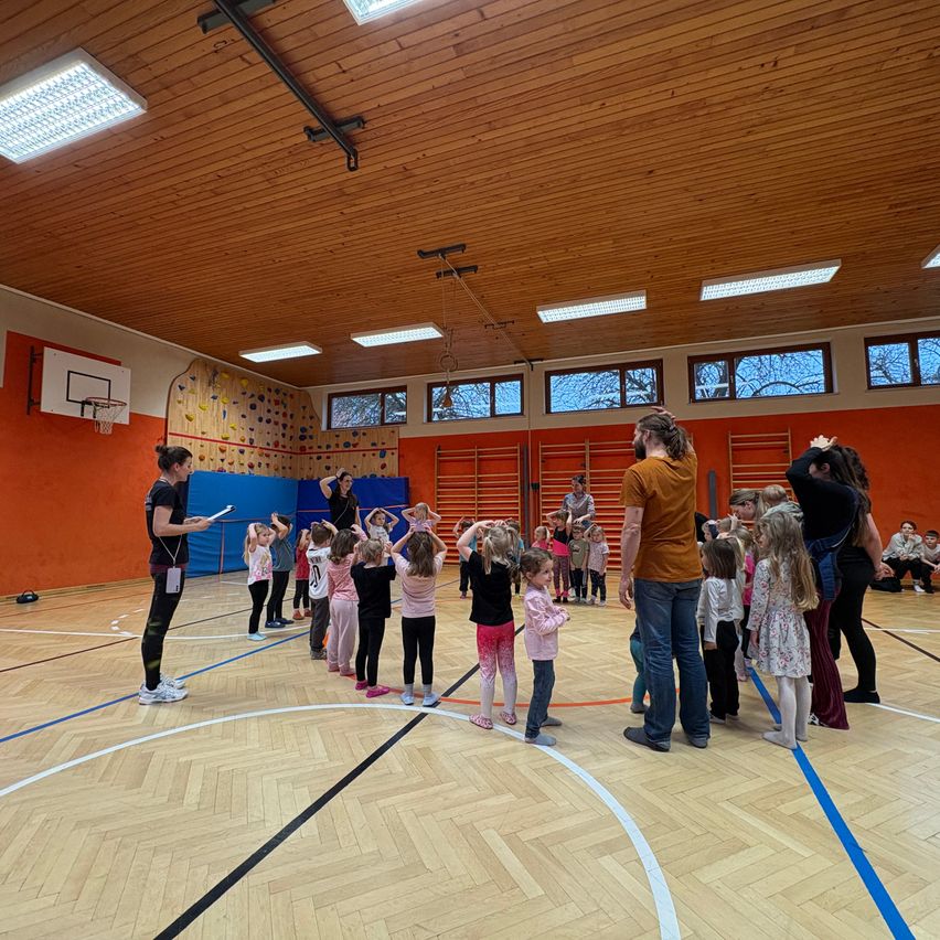 An indoor basketball court with a group of children gathered in a circle. A woman holds a book, likely leading a class. The room has wooden floors, orange walls, and various climbing equipment.