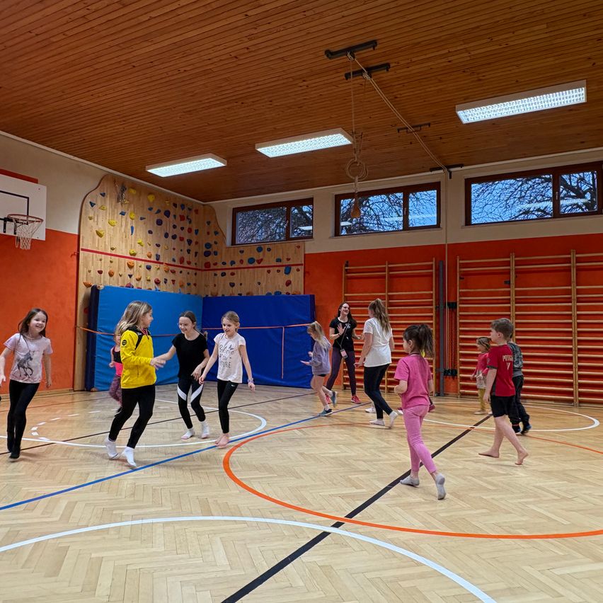 A group of children plays on an indoor court, some with a blue net, under fluorescent lights. A woman is instructing them.
