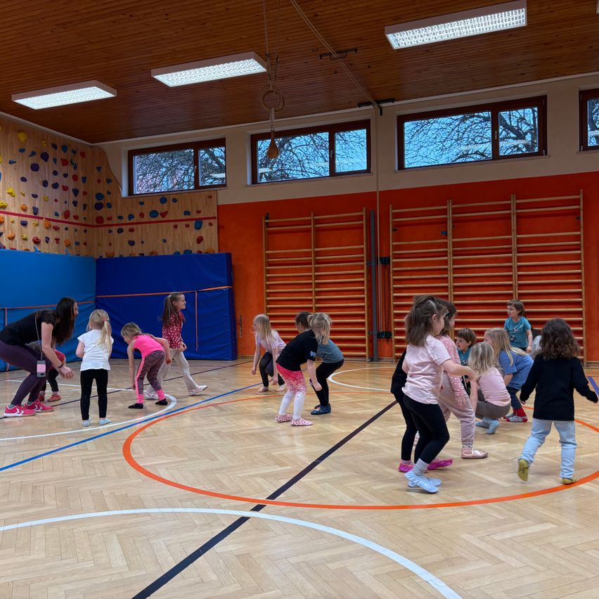 Several children are doing gymnastics in a gym with wooden floors. There are colorful walls and exercise equipment.