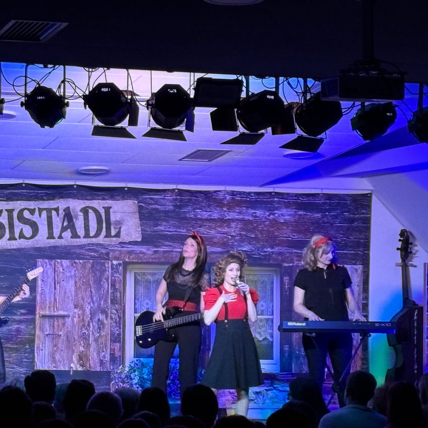 A band is performing on stage under a banner that reads 'Stadl'. Three women are playing instruments, with one singing into a microphone, another playing guitar, and the third playing keyboard. A crowd is watching them.