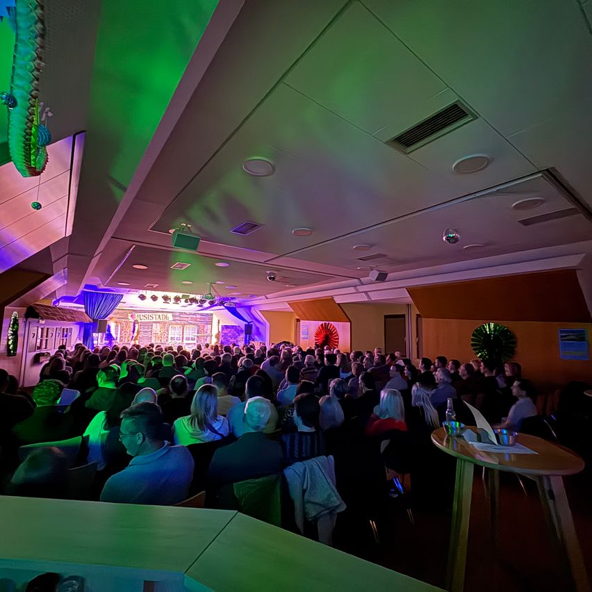 An indoor event with a large crowd, illuminated by green and purple lights, sitting in chairs, with a stage at the front.