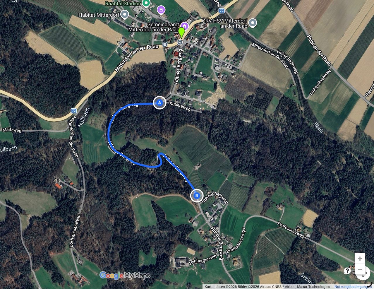 A satellite image of a rural area with various roads, green fields, and buildings. A blue route is marked from point A to point B, passing through several streets.