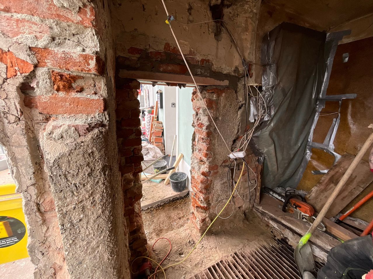A renovation site with exposed brick walls, visible wiring, and a ladder leaning against the wall. Tools and buckets are scattered around.